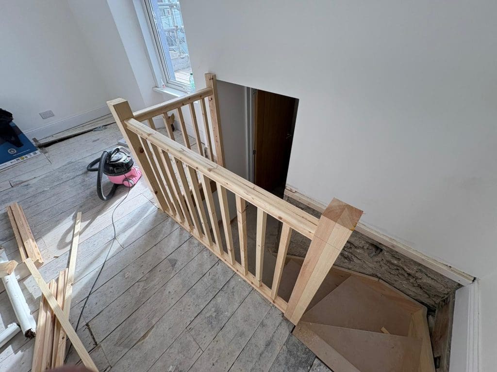 A newly built wooden stair railing surrounds a staircase opening in a room with unfinished wooden floors and white walls. Construction materials and a vacuum cleaner are visible nearby.
