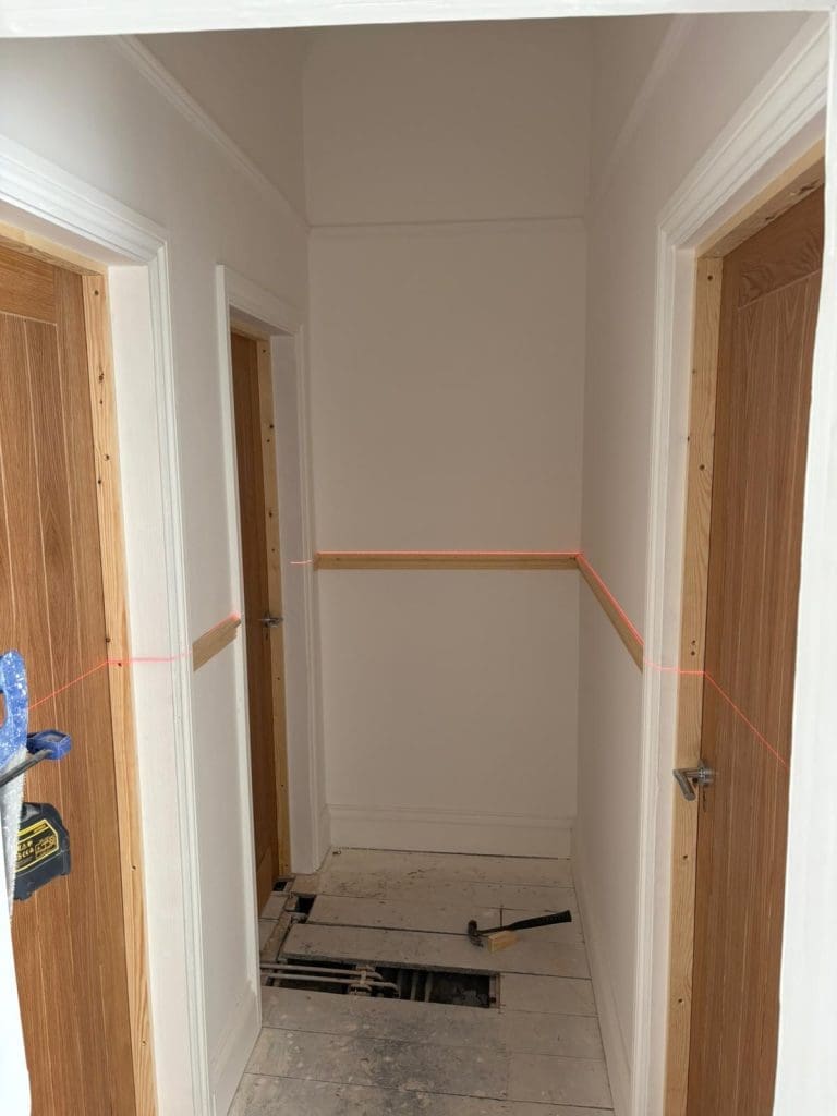 A hallway with white walls and two wooden doors, one on each side. The floorboards are partially removed, exposing openings, and a hammer lies on the floor. A red laser level line runs horizontally across the walls.