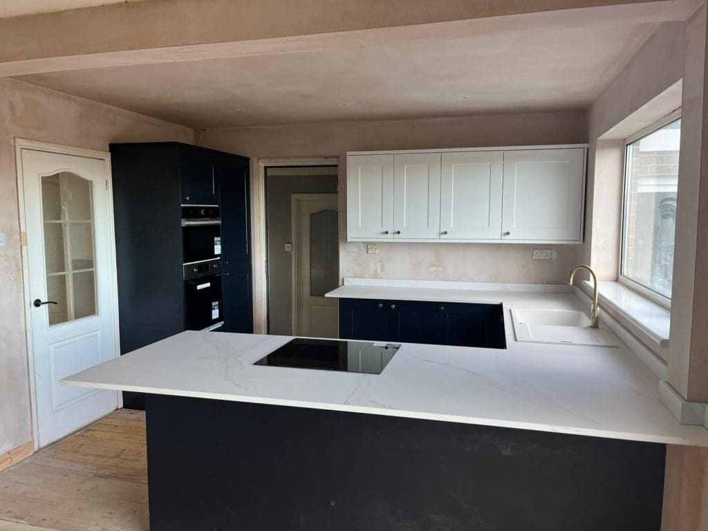Modern kitchen with navy blue lower cabinets, white upper cabinets, a white marble countertop with an integrated stovetop, built-in ovens, a large window, and unfinished plaster walls and wooden floor.
