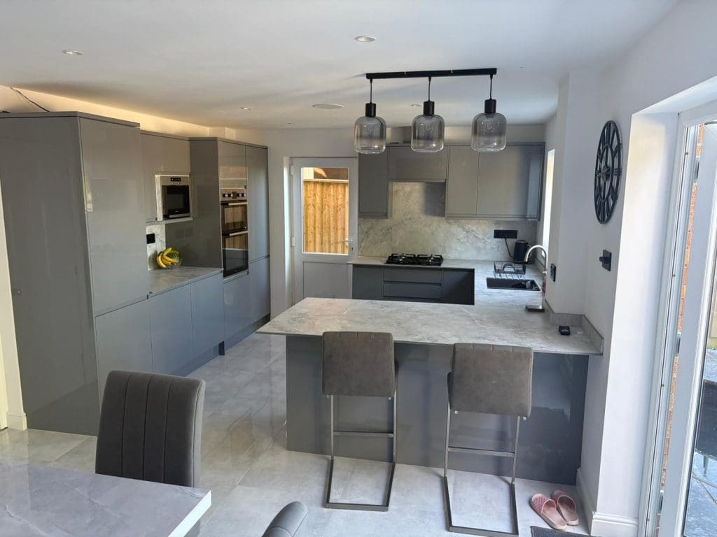 Modern kitchen with grey cabinets, marble countertops, built-in appliances, and an island with two grey bar stools. Three pendant lights hang above the island. A window and sliding glass door bring in natural light.