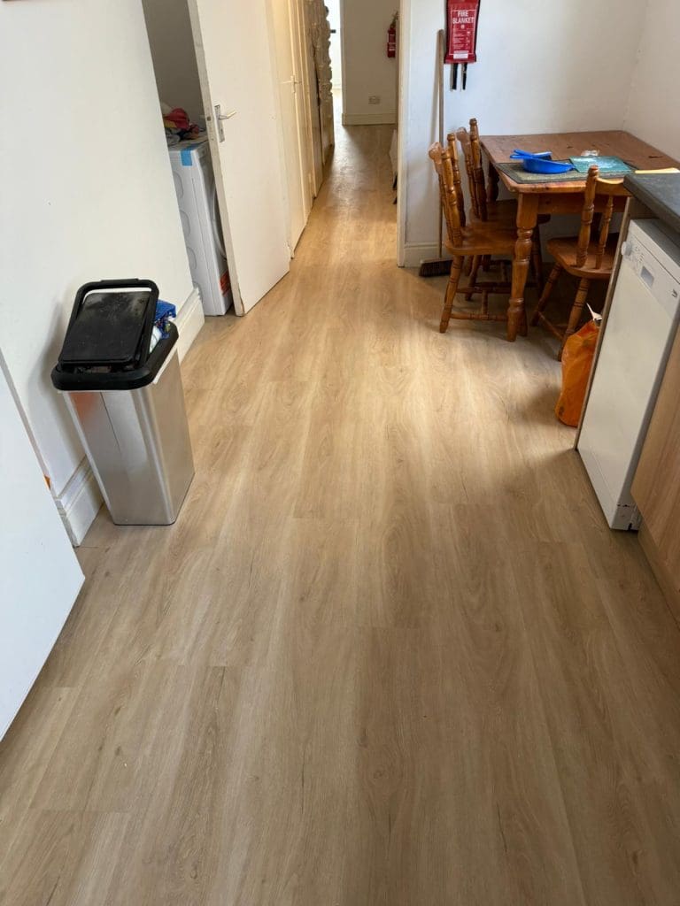 A clean kitchen with light wood flooring, a silver trash bin on the left, a dishwasher and wooden cabinets on the right, and a wooden dining table with chairs in the back near a fire extinguisher on the wall.
