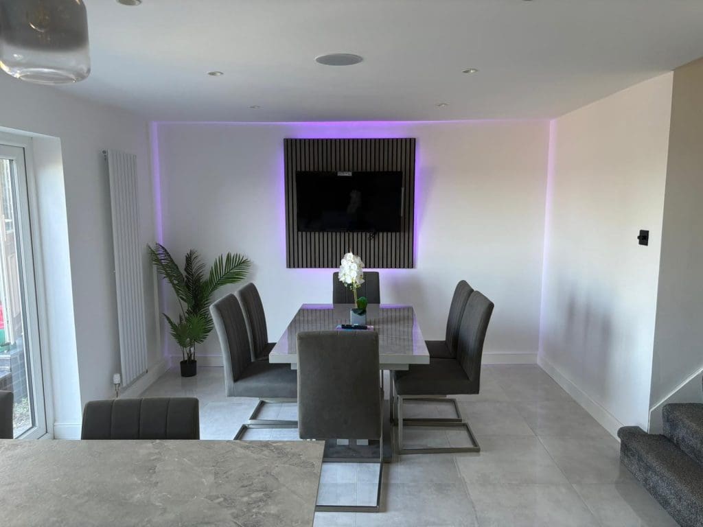 Modern dining room with a rectangular table, six gray chairs, a potted plant, and a wall-mounted TV above a decorative panel. Subtle purple LED lighting accents the wall. Stairs and a glass door are visible.