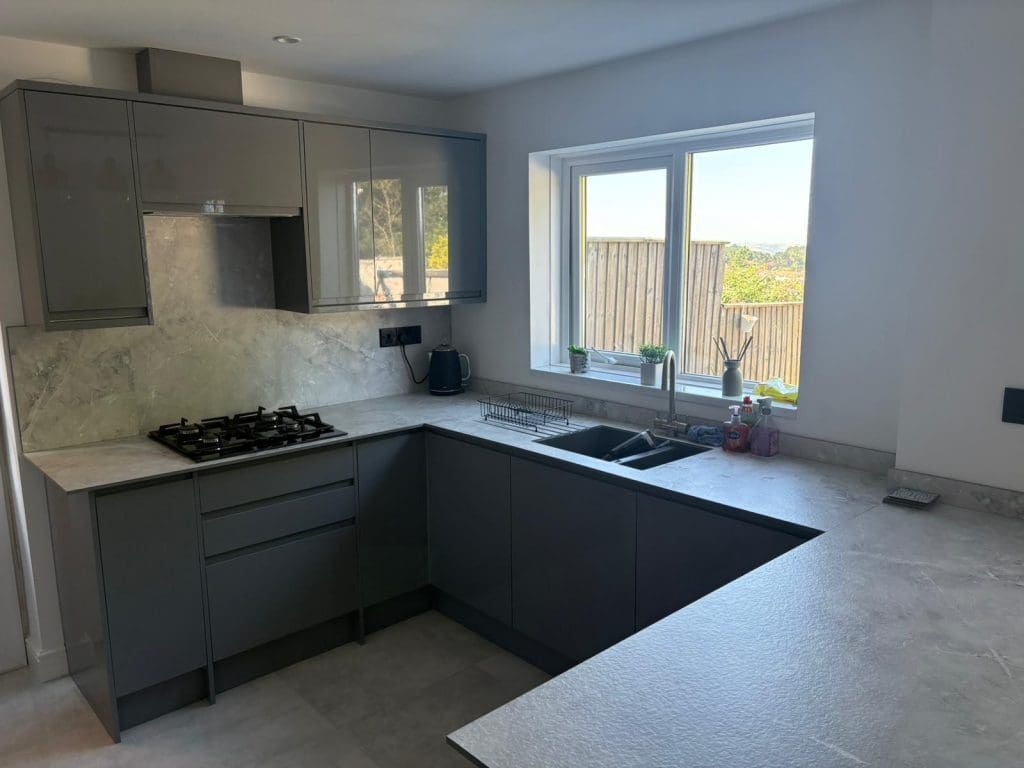Modern kitchen with glossy grey cabinets, a built-in gas stove, and a sunlit window above the sink. Countertops are light-colored, and the space looks clean and uncluttered, with plants and soap by the sink.
