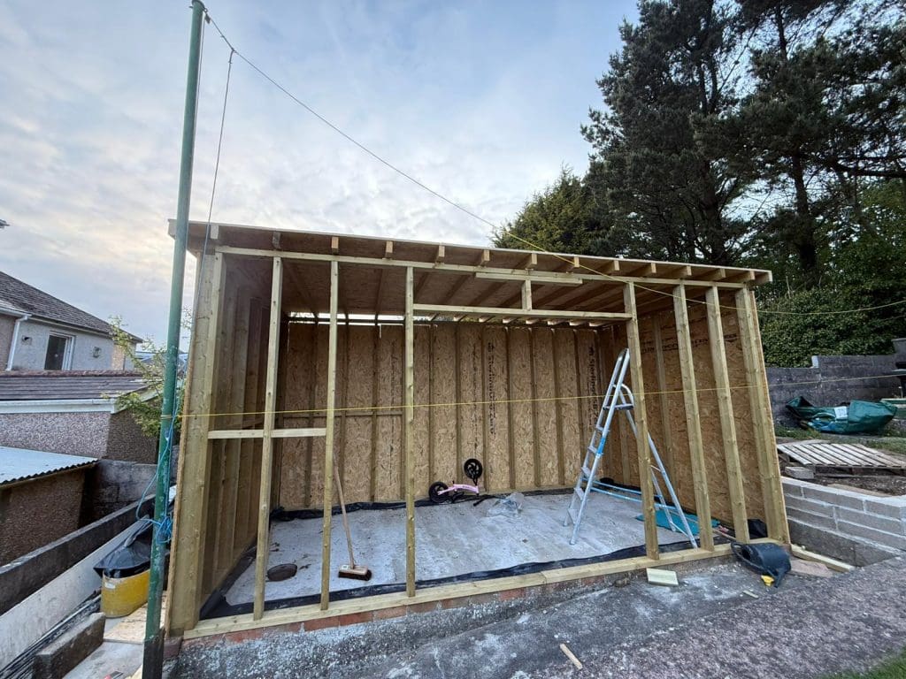 A wooden shed frame under construction in a backyard, with a ladder and construction tools inside. Trees and nearby houses are visible in the background. The roof is partially covered with wood panels.