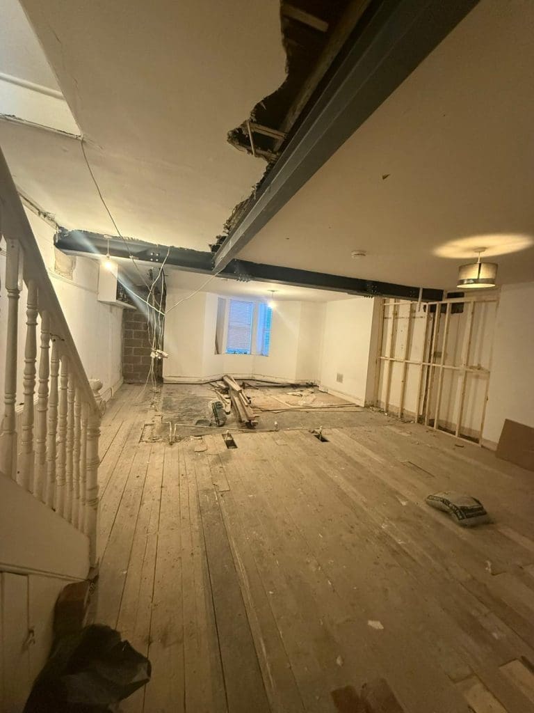 A room under renovation with exposed wooden floorboards, unfinished walls, scattered construction materials, and visible ceiling beams. A staircase is on the left and a window at the back lets in some light.