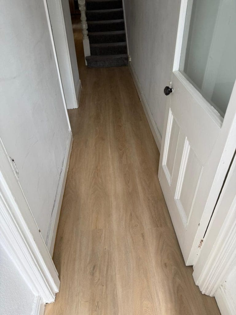 A narrow hallway with light wood flooring, white walls, an open white door on the right, and a staircase with dark carpeting at the end of the hall.
